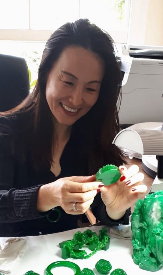 Kehan Li smiling at a microscope desk while examining a piece of imperial green jadeite, surrounded by carved jadeite figures and rough material.